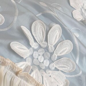 May include: A light blue fabric featuring white floral embroidery with detailed petal stitching. The material has a smooth, slightly reflective surface and includes a gathered, pleated cream-colored trim along the bottom edge and white pearls.