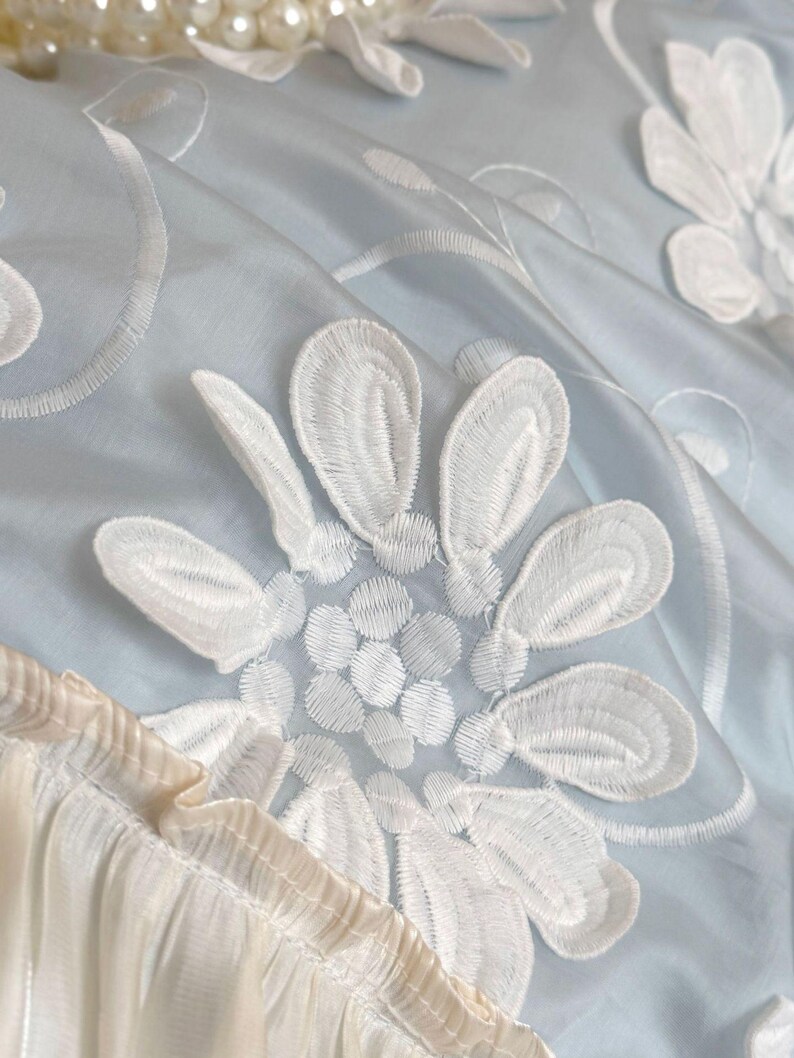 May include: A light blue fabric featuring white floral embroidery with detailed petal stitching. The material has a smooth, slightly reflective surface and includes a gathered, pleated cream-colored trim along the bottom edge and white pearls.