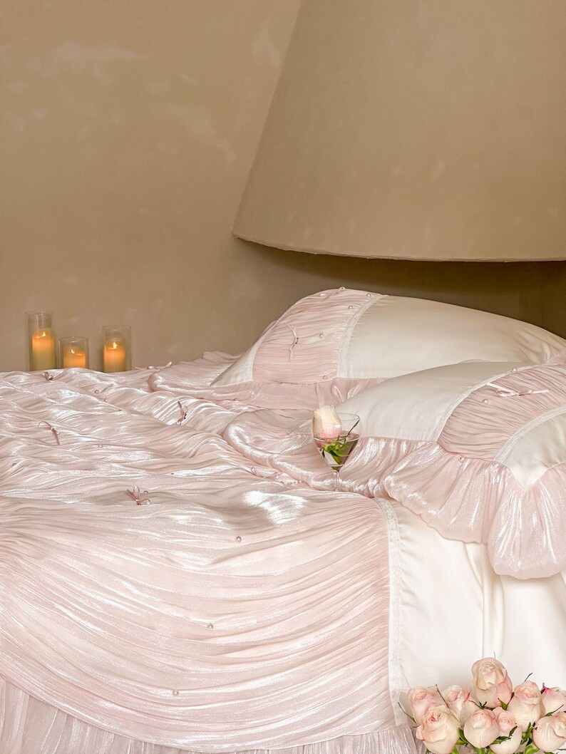 May include: A bedding set featuring a light pink, pleated, and ruffled comforter with small white pearl accents and thin ribbon ties. The set includes matching pillow shams with white centers and pink ruffled edges, accented with small pearls.