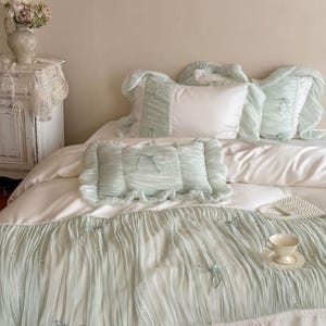 May include: A bedding set featuring a white duvet cover and matching pillowcases with light sage green, pleated, ruffled fabric overlays. The sage green sections are accented with small white faux pearls and thin sage green ribbon bows.
