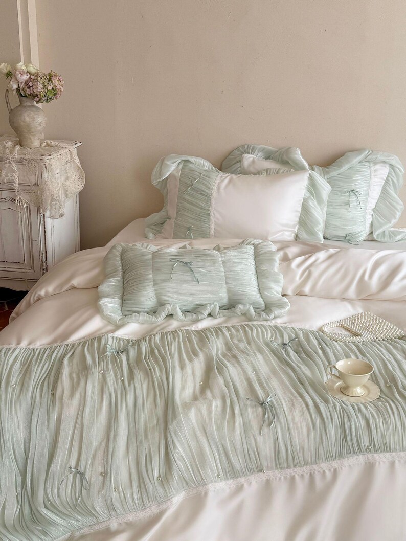 May include: A bedding set featuring a white duvet cover and matching pillowcases with light sage green, pleated, ruffled fabric overlays. The sage green sections are accented with small white faux pearls and thin sage green ribbon bows.