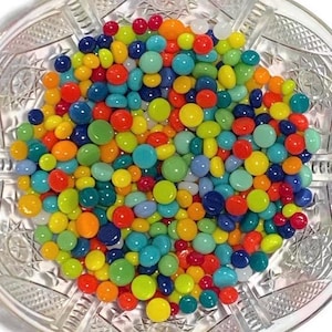 May include: A clear glass bowl filled with colorful, round candies. The candies are in various colors, including red, orange, yellow, green, blue, and white. The bowl has a decorative, etched pattern.