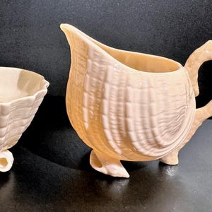 May include: A cream-colored ceramic sugar bowl and creamer set. The bowl is oval with three small feet. The creamer has a handle and spout. Both pieces have a textured, shell-like design.