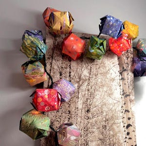 May include: A string of colorful origami paper lanterns in various patterns and colors, including red, orange, green, and blue. The lanterns are strung on a black wire and are displayed against a backdrop of a birch tree forest.