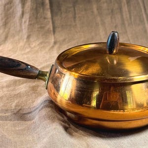 May include: A round copper pot with a polished metallic finish, featuring a long, dark brown wooden handle and a matching lid with a small, dark brown wooden knob centered on top. The pot has a wide, shallow body and a smooth, reflective surface.