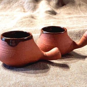 May include: Two terracotta-colored ceramic cups with a rounded body and a single protruding handle. Each cup features a dark brown glazed rim that drips slightly down the exterior, contrasting with the textured, matte finish of the clay body.