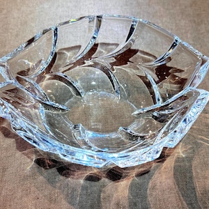 May include: A clear glass bowl featuring a distinct leaf-like pattern with curved, raised ridges radiating from the center toward the scalloped edges. The transparent material reflects light, highlighting the structural design of the glass vessel.