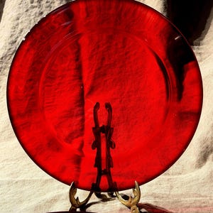 May include: A circular, translucent ruby red glass plate with a smooth, polished surface and a wide, flat rim. The plate is displayed upright on a decorative, ornate gold-toned metal stand that features intricate, curved scrollwork details.