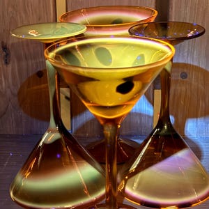 May include: A set of four amber-colored glass martini glasses with a wide, conical bowl and a slender stem. The glass features a distinct gradient effect, transitioning from a deep orange-brown at the base and rim to a lighter, translucent yellow-green center.