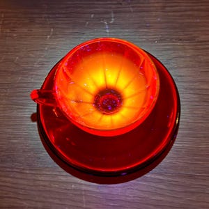 May include: A translucent red glass teacup and matching saucer set. The cup features a ribbed interior design that radiates from the center, creating a glowing orange and yellow light effect within the bowl of the cup when illuminated from above.
