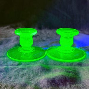 May include: A pair of matching candle holders made of translucent, vibrant neon green glass. Each piece features a wide, circular base that tapers upward into a smooth, rounded stem, topped with a small, circular opening for a candle.