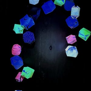 May include: A string of decorative paper lanterns shaped like folded cubes, featuring a variety of colors including blue, green, pink, and white, with some displaying polka dot patterns and others showing solid or textured surfaces.