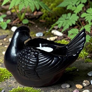 May include: A black ceramic dove-shaped candle holder with a white candle inside. The dove has detailed wing and feather textures. The candle holder is set on a stone surface with moss and small rocks, with green foliage in the background.
