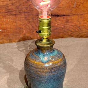 May include: A table lamp featuring a rounded, glazed ceramic base with mottled blue and brown tones. The lamp has a brass-colored metal socket with a black push-button switch and a clear, round glass light bulb with a visible glowing red filament.