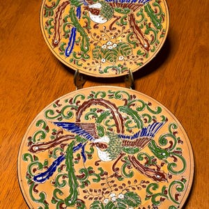 May include: Two round ceramic plates featuring a vibrant orange background with a central bird motif in shades of blue, green, and brown. The design includes intricate swirling foliage, leafy branches, and small white floral accents across the surface.
