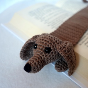 May include: A brown crocheted dachshund bookmark with black eyes and a black nose. The bookmark is lying on a book.