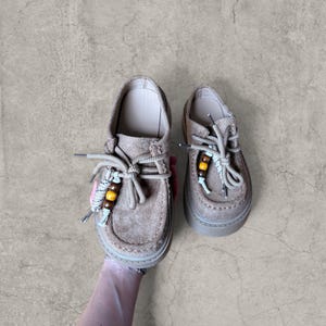 Op de afbeelding: Een paar bruin suède schoenen met dikke zolen. De schoenen hebben beige veters met decoratieve houten kralen. De schoenen worden door een hand vastgehouden.