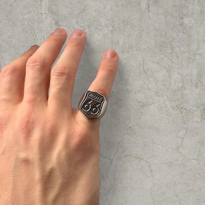 May include: A silver-colored ring featuring the Route 66 shield design. The ring has a rectangular face with the words "ROUTE 66" and the number "66" in a vintage style. The ring is worn on a finger.