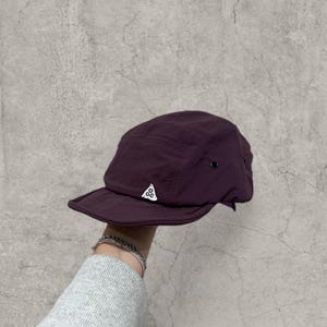 Peut inclure: Casquette violet foncé avec une petite visière et un logo blanc. Fabriquée dans un matériau lisse, possiblement en nylon, elle présente un design structuré à cinq panneaux et un petit bouton noir sur le côté.