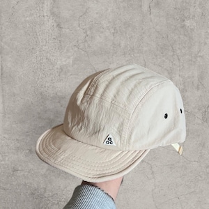 May include: A beige five-panel cap with a curved peak. The cap has a small logo on the side and two small black eyelets. The hat is made of a lightweight material and has a structured design.