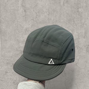 Peut inclure: Casquette vert foncé à cinq panneaux avec une petite visière. La casquette présente un petit logo triangulaire blanc à l'avant. Fabriquée dans un matériau durable et léger, elle a une conception structurée.