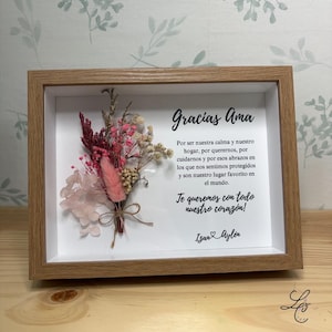 Puede incluir: Un arreglo floral enmarcado con un marco de madera clara. El arreglo incluye flores secas en tonos rosa, rojo y crema, atadas con hilo. El texto en español dice "Gracias Ama" y un mensaje de amor.