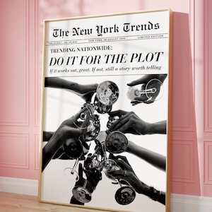 May include: Black and white framed print featuring a newspaper headline that reads "The New York Trends." The headline "TRENDING NATIONWIDE: DO IT FOR THE PLOT" is displayed above a photo of hands toasting with wine glasses. The print is displayed on a pink wall.