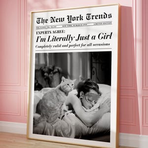 May include: A framed black and white print featuring a vintage newspaper headline, "The New York Trends." The headline reads "I'm Literally Just a Girl." The image shows a person wearing an eye mask in bed with a cat.
