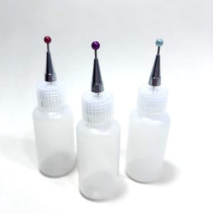 May include: Three translucent plastic squeeze bottles with fine metal precision tips. Each bottle features a screw-on cap and a small, coloured ball-topped pin inserted into the tip, with one red, one purple, and one light blue ball topper.