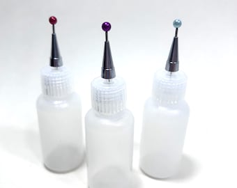 May include: Three translucent plastic squeeze bottles with fine metal precision tips. Each bottle features a screw-on cap and a small, colored ball-topped pin inserted into the tip, with one red, one purple, and one light blue ball topper.