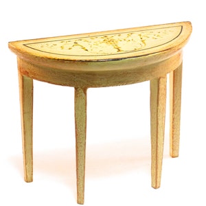 Puede incluir: Una mesa de madera de media luna pintada de amarillo con un diseño floral decorativo en la parte superior. La mesa tiene tres patas.