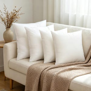 May include: Four square white throw pillows arranged in a row on a cream-colored sofa. A textured tan throw blanket with a geometric chevron pattern is draped over the front of the sofa, and a beige vase with dried flowers sits to the side.
