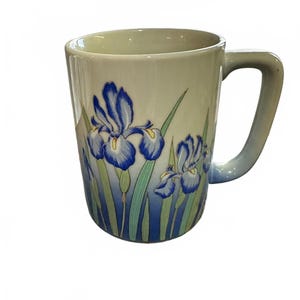 Puede incluir: Una taza de cerámica con una base de color crema que se transforma en un azul profundo en la parte inferior. La taza presenta un diseño pintado de flores de iris azules con centros amarillos y tallos y hojas verdes. Tiene una asa lateral.