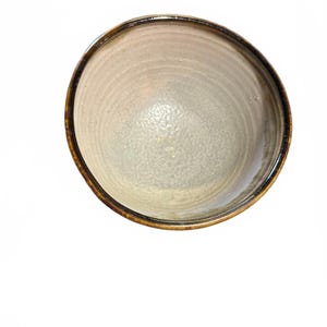 May include: A round ceramic bowl with a speckled off-white interior and a smooth, glossy finish. The rim is defined by a dark brown, toasted edge that contrasts with the lighter center, showing subtle concentric rings from the pottery process.