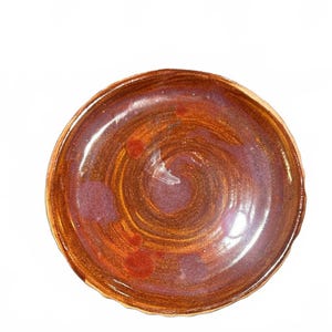May include: A round ceramic dish with a glossy finish, featuring a swirling pattern of deep copper, bronze, and reddish-brown tones. The surface has subtle variations in color and texture, with a slightly uneven, scalloped edge along the rim.