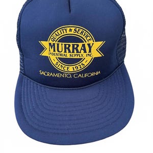 May include: A navy blue trucker hat with a mesh back and a curved brim. The front of the hat features a yellow circular logo with the words "QUALITY & SERVICE" and "MURRAY INDUSTRIAL SUPPLY, INC. SINCE 1925 SACRAMENTO, CALIFORNIA."