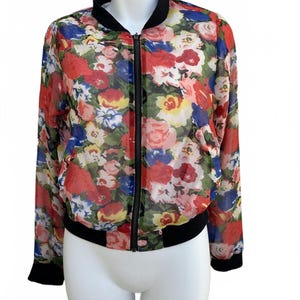 May include: A floral print bomber jacket with a black zipper, collar, and cuffs. The jacket features a vibrant pattern of red, pink, blue, and yellow flowers on a sheer fabric. The jacket is displayed on a white mannequin.