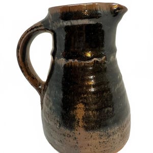 May include: A ceramic pitcher with a glossy finish, featuring a dark brown and black glaze that transitions into a lighter, speckled tan base. The vessel has a curved handle on the side and a small pouring spout at the top rim.