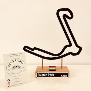 以下が含まれることがあります： 木製の台座に取り付けられた、Balaton Parkレーストラックの黒いアウトライン。ベースにはトラック名と距離4.115kmが含まれています。小さなハンガリーの旗も付いています。