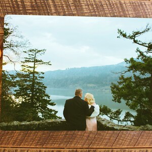 May include: A couple stands on a cliff overlooking a lake and mountains. The man is wearing a black suit and the woman is wearing a pink dress. The sky is blue and there are trees in the foreground.
