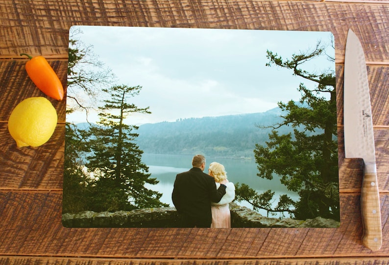 May include: A couple stands on a cliff overlooking a lake and mountains. The man is wearing a black suit and the woman is wearing a pink dress. The sky is blue and there are trees in the foreground.