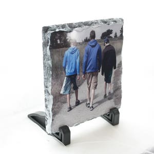 May include: A photo slate with a printed image of three people walking on a path. The people are wearing casual clothing and are walking away from the camera. The slate is on a black stand.