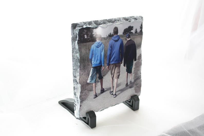May include: A photo slate with a printed image of three people walking on a path. The people are wearing casual clothing and are walking away from the camera. The slate is on a black stand.