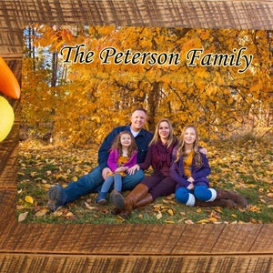 May include: A family of four poses in front of a tree with yellow leaves. The parents are sitting on the ground with their two daughters. The parents are wearing casual clothing and the daughters are wearing dresses. The text "The Peterson Family" is written at the top of the image.