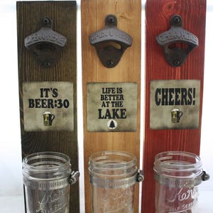 May include: Three wooden bottle opener wall mounts with mason jar holders. The bottle openers are black metal and the mason jars are clear glass. The wood is stained in different colours: dark brown, light brown, and red. The signs on the wood read "It's Beer:30", "Life is Better at the Lake", and "Cheers!"