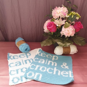 Puede incluir: Dos paños de cocina de ganchillo con el texto "Keep Calm & Crochet On" en hilo blanco sobre fondo azul y blanco. Los paños de cocina están sobre una superficie rosa con dos ovillos de hilo y un ramo de flores rosas en el fondo.
