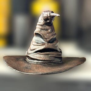 Può includere: Un cappello parlante marrone dettagliato, con tesa larga e punta. Il cappello ha un aspetto invecchiato con pieghe visibili e una bocca leggermente aperta. Replica del Cappello Parlante della serie di Harry Potter.