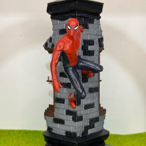 Op de afbeelding: Een Spider-Man beeldje in een dynamische pose, vastklampend aan een bakstenen structuur. De figuur is overwegend rood en zwart, met een gedetailleerd masker en pak. De bakstenen structuur is grijs en zwart, met een zwarte basis en bovenkant. Het beeldje staat tegen een groene achtergrond.