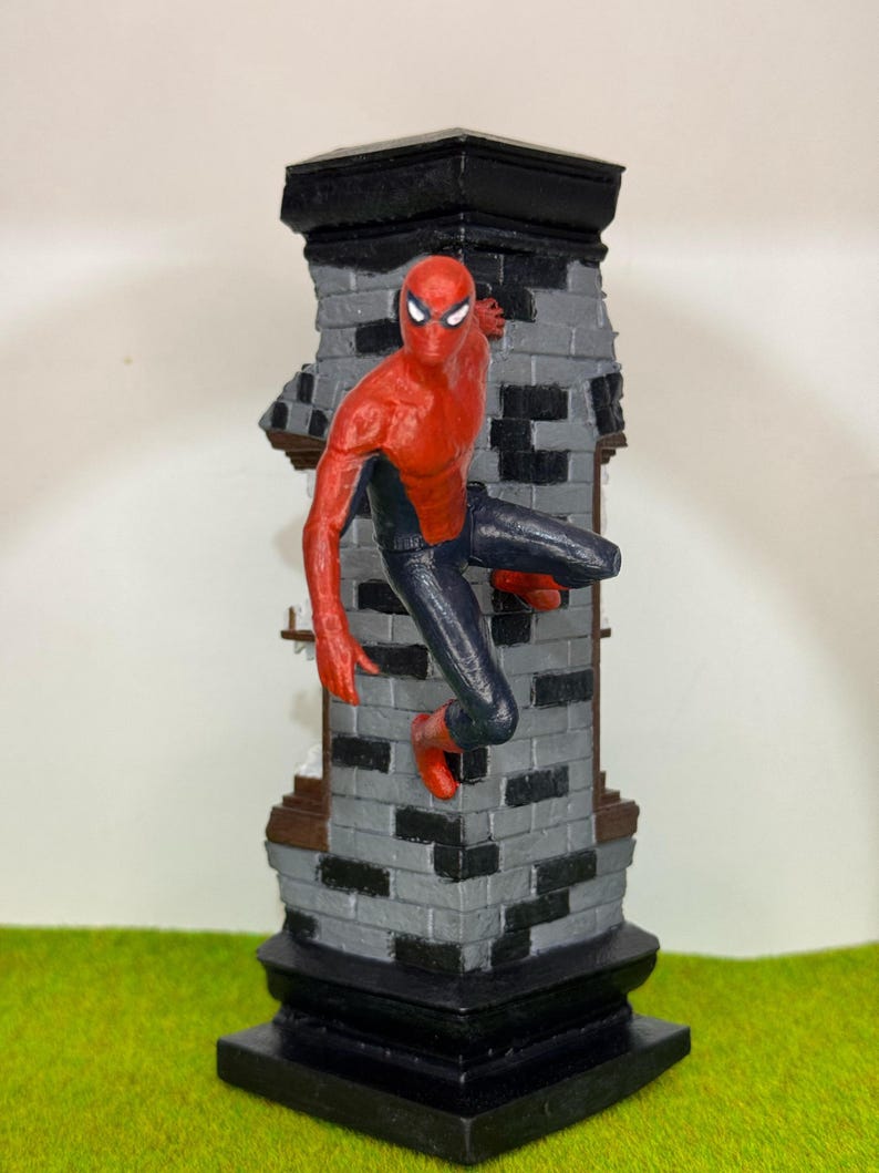 Op de afbeelding: Een Spider-Man beeldje in een dynamische pose, vastklampend aan een bakstenen structuur. De figuur is overwegend rood en zwart, met een gedetailleerd masker en pak. De bakstenen structuur is grijs en zwart, met een zwarte basis en bovenkant. Het beeldje staat tegen een groene achtergrond.