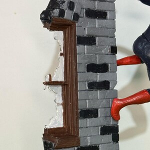 Op de afbeelding: Gedetailleerd beeldje van een bakstenen muur met een gebroken raam. De muur is grijs en zwart geschilderd, met een bruin kozijn. Een figuur in een rood en blauw pak beklimt de muur.